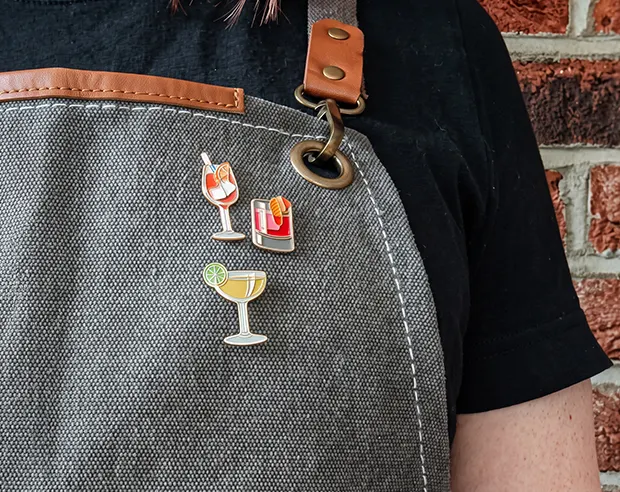 Custom cocktail enamel pins displayed on a grey barista apron