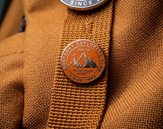 A round orange "Monterey Explorers Club" mountain pin on an earthy yellow canvas backpack strap