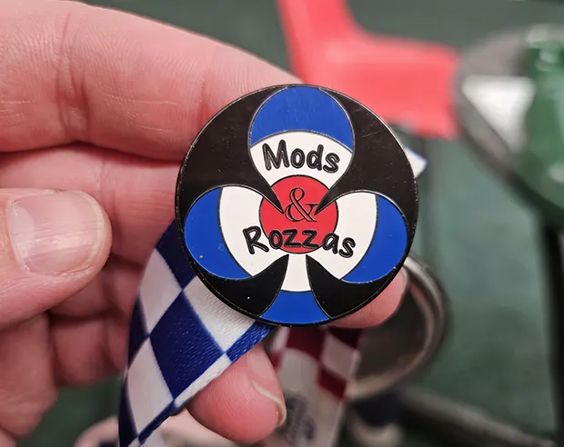 A circular “Mods & Rozzas” hard enamel pin on a lanyard ribbon, held in hand