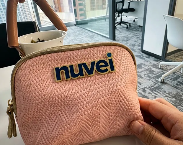 A custom "nuvei" logo pin on a pink woven makeup handbag