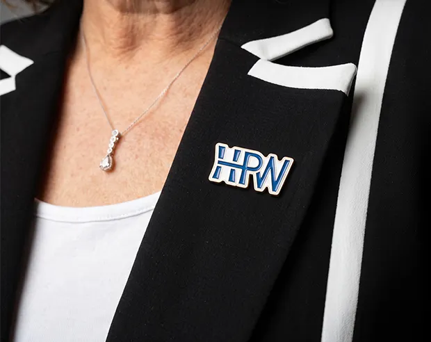 A custom blue "HPW" letter enamel pin on a black and white suit lapel