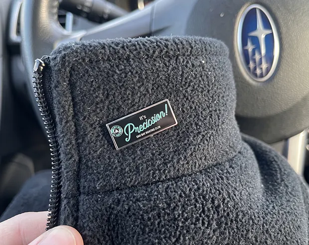A "It's Precision!" rectangular hard enamel pin on the collar of a fleece sweatshirt jacket