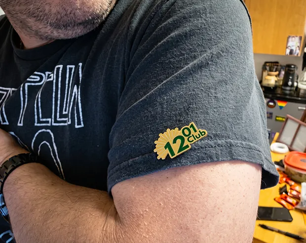 A gold plated green "1201 Club" pin on the sleeve of a dark grey t-shirt