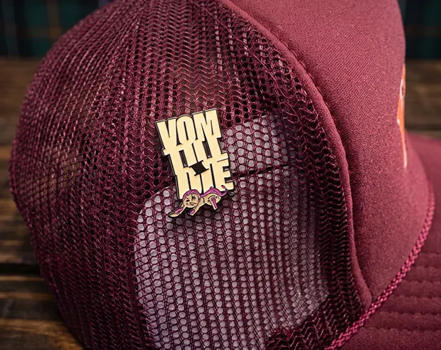 A custom typography enamel pin displayed on the side of a burgundy trucker hat.